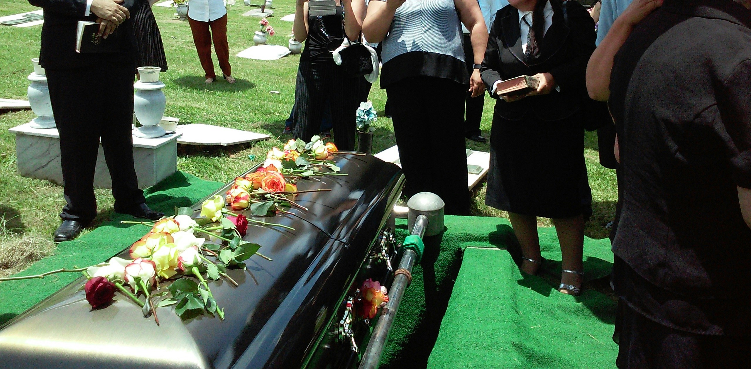 People stand in a cemetery beside a casket with flowers laid across the top.