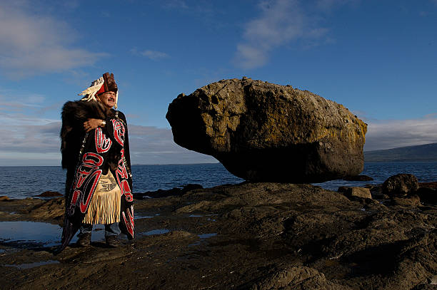A Haida Gwaii man dressed in traditional regalia stands on a beach.