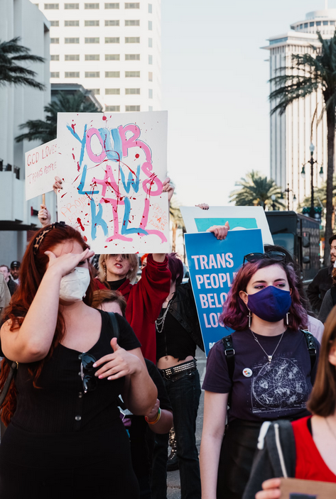 Protesters hold up a sign that says "Your laws kill" in the colors of the transgender pride flag.