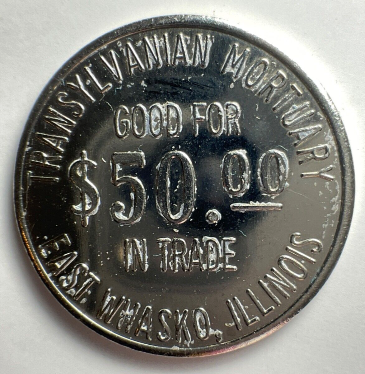 A silver colored embalmers token with the words "Transylvanian Mortuary, East Whasko, Illionois, Good For Fifty Dollars in Trade"