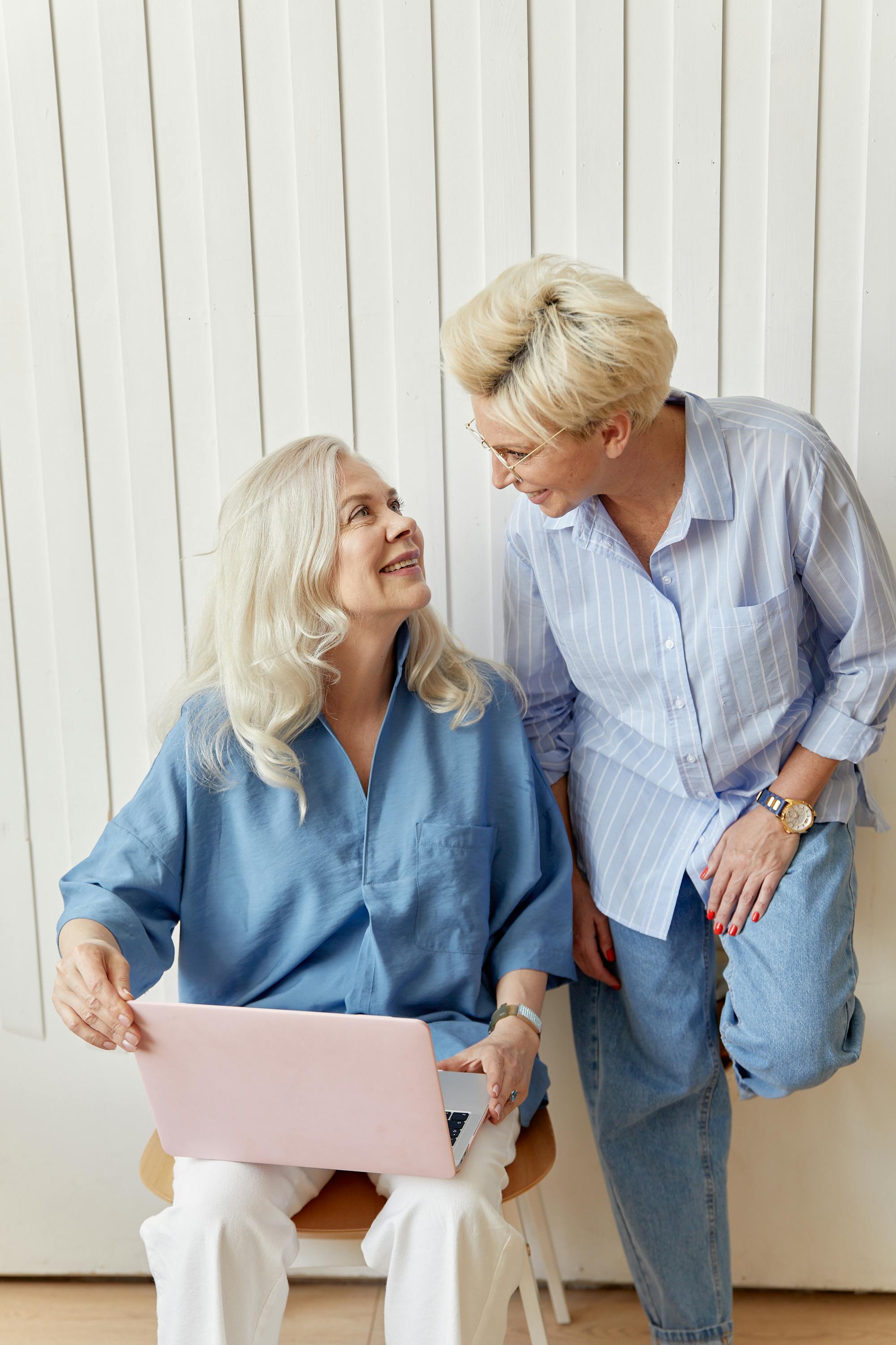 Two older women, one seated with a laptop and one standing, smile at eachother.